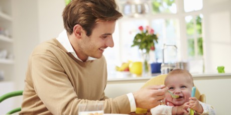 Father Feeding Baby Sitting In High Chair At Mealtime